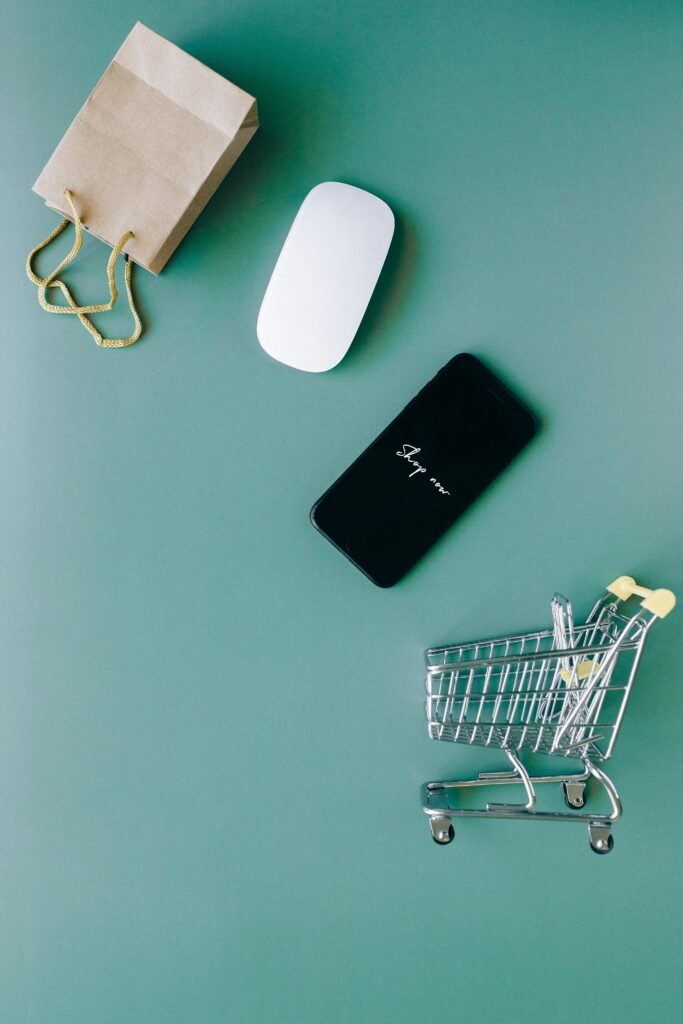 Top view of a smartphone, mouse, paper bag, and mini shopping cart on a green background, symbolizing e-commerce.